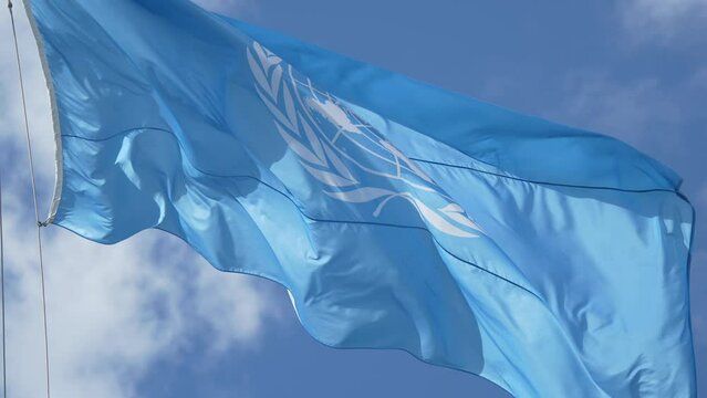 4K video of the United Nations (UN) organisation flag winding against a blue sky with white clouds, symbol image for peace around the globe. New York, UN Headquarters, 2022.