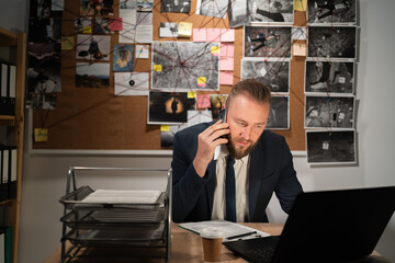 young detective sitting at police department talking to colleague on mobile phone, evidence board on background. Portrait of cop having phone call with witness in office