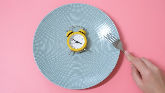 Female hand holding an alarm clock on background of empty plate, concept of intermittent fasting.
