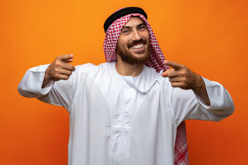 Young Arab man in traditional clothing pointing on you on orange background