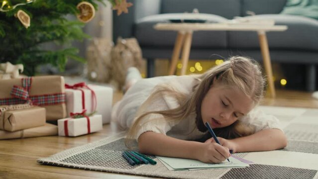 Little caucasian girl lying on carpet next to Christmas tree and writing a letter to Santa Claus. Shot with RED helium camera in 8K.   
