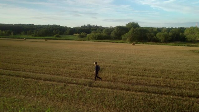 Man traveling alone with a backpack through a deserted field landscape after harvest. Straw bales, trees and evening blue sky in the background. Romantic quiet nature to relax