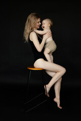 Vertical portrait of young blonde barefoot woman in black bodysuit embracing and playing with toddler baby on black view