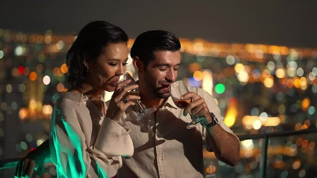 4K Caucasian couple celebrating holiday event at luxury skyscraper rooftop bar at night. People enjoy city nightlife having dinner and drinking wine at outdoor rooftop restaurant on summer vacation.