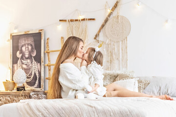 Mom kisses her little daughter sitting on the bed in a bright bedroom.Family. Ethno-interior design