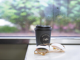 Black paper cup of coffee on vintage table over window glass with rain drops