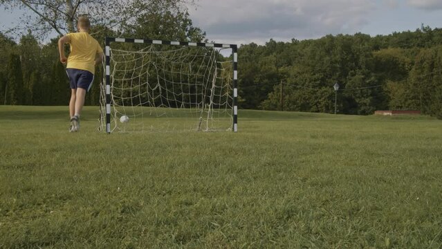 A young boy kicks the ball into the goal, but there is a tear in the net of the goal, so the ball rolls through the net. Location: Hungary, Nagybörzsőny mountains.