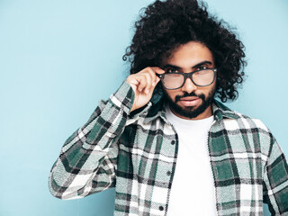 Handsome smiling hipster  model. Sexy unshaven Arabian man dressed in summer shirt and jeans clothes. Fashion male with long curly hairstyle posing near blue wall in studio. In spectacles, glasses