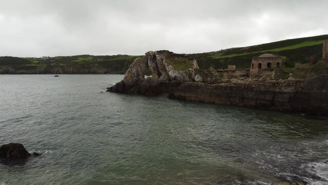 Reverse aerial view flying low towards Traeth Porth Wen Beach bay abandoned brickwork site on the Irish sea coast