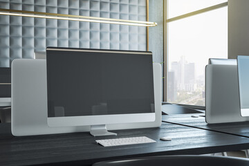 Black blank modern computer monitor screen with place for your logo or text on dark wooden table in sunlit coworking office with city view and golden lamps above workspaces. 3D rendering, mock up