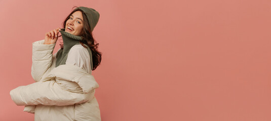 Cheerful young caucasian girl is smiling looking into camera on pink background with place for text. Dark-haired lady wears grey hat, sweater, and white jacket. Lifestyle, feminine beauty concept