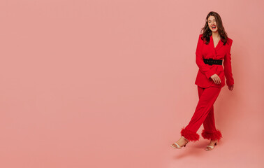 Fototapeta na wymiar Full-length young caucasian girl in red pantsuit poses merrily against pink background. Brunette with wavy hair laughs looking into camera. People's emotions, lifestyle and fashion concept. 