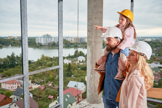 Daughter Sitting On Father's Shoulders And Pointing At Something At Construction Site. Family With Child Future Homeowners Observing Apartment Building Under Construction.