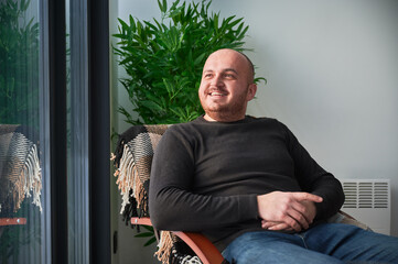 Smiling man enjoying weekends inside contemporary barn house. Happy male tourist sitting on chair in new cottage.