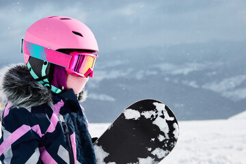 Little girl snowboarder with equipment helmet and goggles outwear holding snowboard resting on top...