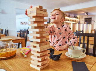 Kid girl playing stacking wood blocks (Jenga) funny and joyful on game in cosiness coffee house. Hand movement control building computational skills children's play concept.