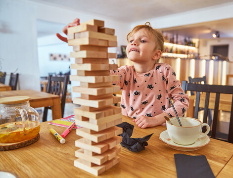 Kid girl playing stacking wood blocks (Jenga) funny and joyful on game in cosiness coffee house. Hand movement control building computational skills children's play concept.