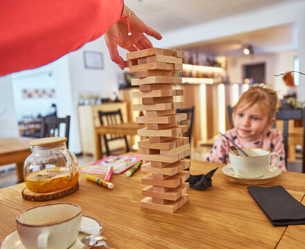 Kid girl playing stacking wood blocks (Jenga) funny and joyful on game in cosiness coffee house. Hand movement control building computational skills children's play concept.