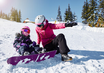 Fototapeta na wymiar Mother teaches his daughter to snowboard. Woman sits and showing way to ride on ski slope at sunny winter day, travel vacation, landscape mountains background.