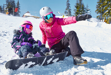 Fototapeta na wymiar Mother teaches his daughter to snowboard. Woman sits and showing way to ride on ski slope at sunny winter day, travel vacation, landscape mountains background.
