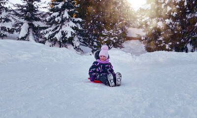 Fototapeta na wymiar Little girl enjoying the winter sledding time. Child playing and having fun riding on a snowy hill. Slide down from snow slope sitting in plate