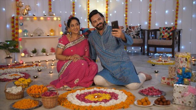 An attractive husband-wife in traditional attire clicking pictures - Diwali celebrations  Hindu festival. An Indian couple sitting on the floor - colorful lighting and decoration  Diwali riutals  f...