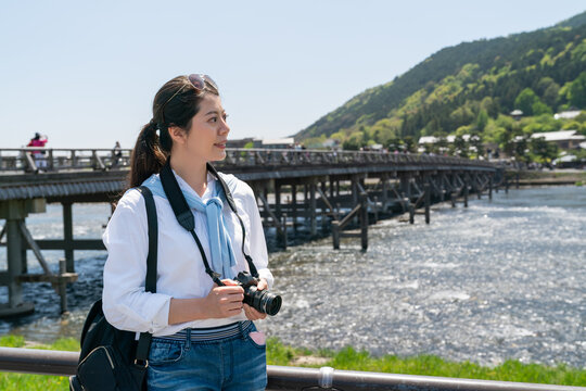 smiling asian Taiwanese woman traveler enjoying nice landscape by at riverside of Katsura River with Togetsu-kyo Bridge at background hile traveling to arashiyama in Kyoto japan on sunny day