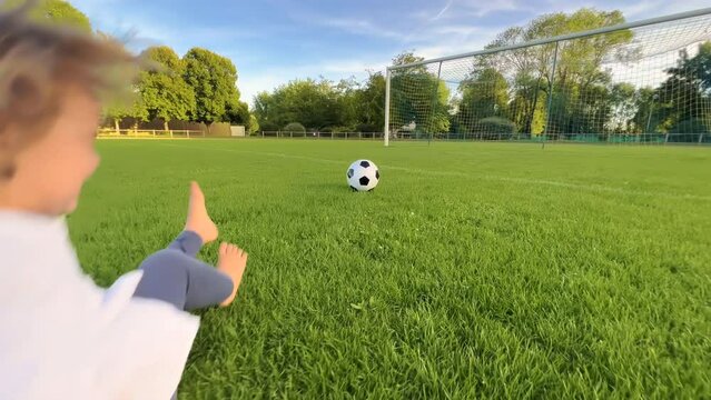 Little kid plays football at sunset, baby runs around green football field and kicks ball, girl runs to floor in meadow and smiles, learns how to kick ball, dream of becoming team footballer
