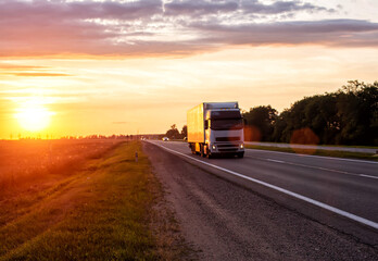 A truck with a trailer transports cargo in the summer against the backdrop of sunset and beautiful evening nature. The concept of logistics problems in trucking, 
