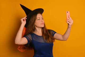 Young witch girl taking selfie photo on cellphone or smartphone and holding black hat on a yellow background. Wizard woman doing selfie shot on mobile cell phone and wearing blue dress.