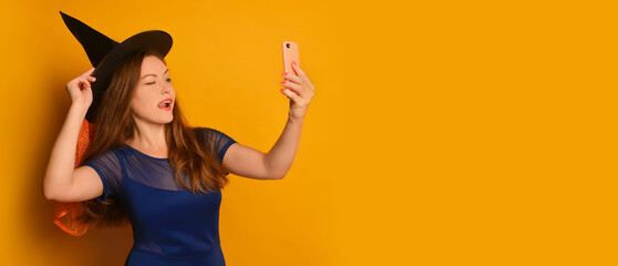 Young witch girl taking selfie photo on cellphone or smartphone and holding black hat on a yellow background.

Halloween woman doing selfie shot on mobile phone and sticking tongue out in blue dress.