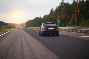 Fototapeta na wymiar A black passenger car drives along an asphalt highway in the evening against the background of a sunset. The concept of traffic on the road and travel in your car. Copy space for text