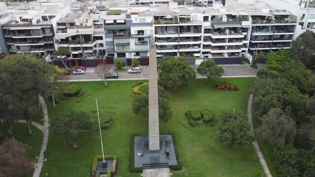 4k drone video of a park with a obelisc. Drone flies in orbit around the obelisc monument from left to right. Apartment buildings next to the park. Located in Lima, Peru in San Borja district.