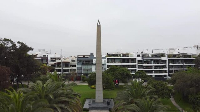 4k video of a drone lifting off and climbing altitude in a park with a tall obelisc monument, and many trees and green grass, behind it apartment buildings. Located in Lima, Peru in San Borja district