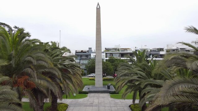 4k drone video of a park with a obelisc. Drone flies in between palm tree tops towards the obelisk. Behind it apartment buildings, and behind a gray sky. Located in Lima, Peru in San Borja district.