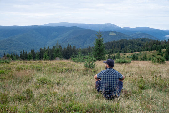 Hiker Beard Man Is Sitting In The Grass And Looking At The Mountain View. Dressed Up In A Cap, Shorts, And Plaid Shirt With