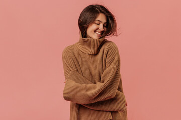 Fototapeta premium Smiling young caucasian girl looking away with her arms around herself in pink background. Dark-haired lady is wrapped in brown sweater. Mood, lifestyle, concept .