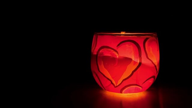 Glass candlestick. Burning candle on a black background. Romantic atmosphere for Valentines Day. Decorative red pink candle with a heart pattern. Side view. Slow motion. Copy space. Love divination.