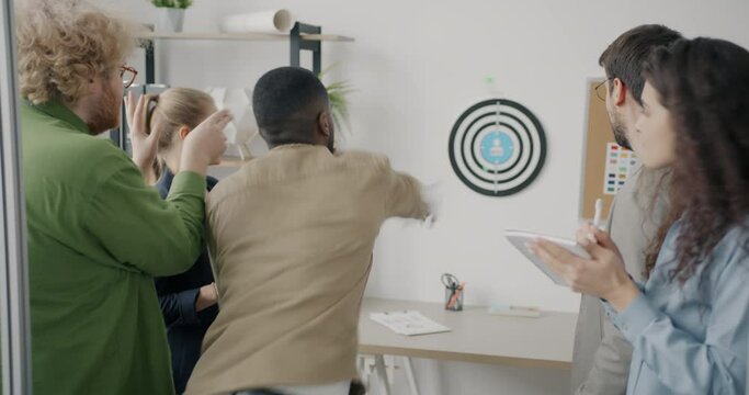 African American man is playing darts at work while colleagues diverse group are watching and cheering. Workplace fun and youth concept.