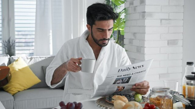 Young man in bathrobe enjoying slowly brakfast, coffee and newspaper.