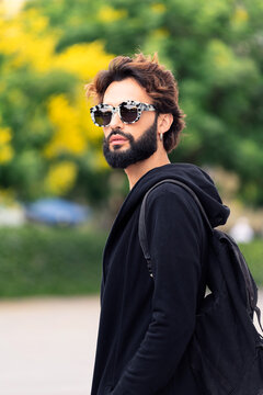 portrait of a beautiful young man with backpack and fashionable sunglasses walking by a city park, concept of urban lifestyle and stylish clothing, copy space for text