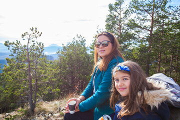 Portrait of smiling family on the peak of the mountain. Mother and daughter sitting on a bench, viewing a beautiful autumn nature landscape. Active people lifestyle.