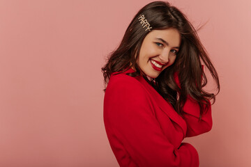Naklejka premium Attractive young caucasian girl with toothy smile on her face looks at camera on pink background. Brunette with loose hair is wearing red elegant jacket. People's emotions, fashion concept. 