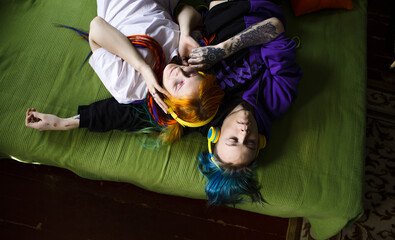 Fototapeta na wymiar Portrait of a young tattooed punk couple of a girl and a guy with long dyed hair braided, lying on a large green bed, top view. They listen to music. Yellow large headphones. Stylish modern youth