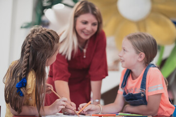 Creative kids during an art class in a daycare center or elementary school classroom drawing with female teacher. 