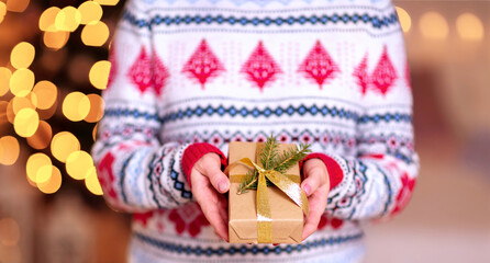 Fototapeta na wymiar Elderly Female hands hold out a gift wrapped in kraft paper and decorated with a live spruce twig and a gold shiny ribbon on a background of a sweater with Christmas ornaments and festive illumination