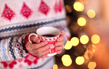 Close-up female hands with neat manicure hold a tea mug decorated with Christmas ornaments on the background of New Year's lights