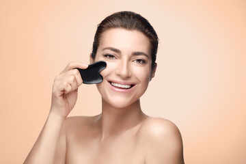 Beauty anti aging and skin care concept. Beautiful middle aged woman with fresh clean skin doing a lifting massage with a Gua sha tool. Beige background with copy space