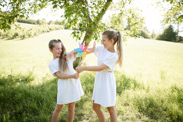 Fototapeta premium two pretty girls, sisters with white dresses holding colorful pinwheels and standing in green meadow in front of a walnut tree and are happy