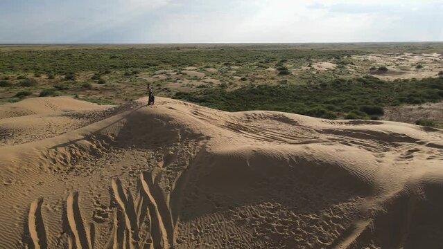 drone is circling around a girl in a long leopard dress stands on a high dune in a desert with grass in summer in Astrakhan